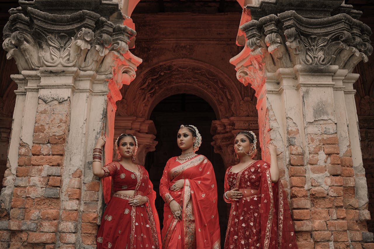digital Three women in vibrant red sarees pose elegantly by ancient architecture.